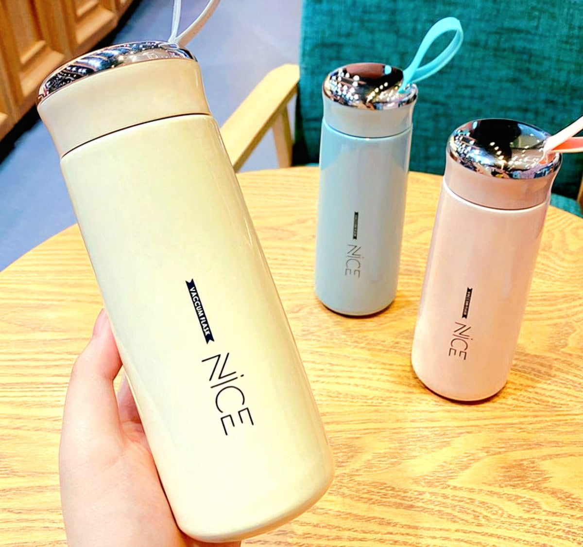 A hand holds a large cream-colored insulated water bottle with a metallic patterned lid and the brand name 'NICE' printed vertically. Two smaller insulated bottles, one light blue and one light pink, sit on a wooden table in the background.