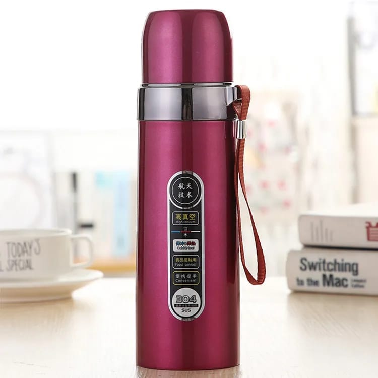 A metallic magenta pink vacuum insulated water bottle with a brown carrying strap, standing on a light wooden table with a blurred coffee cup and books in the background.