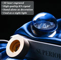 A blue glass crystal ball with a 3D laser-engraved solar system design, displayed on a dark book with a wooden socket and draped blue fabric. A text overlay on the top left lists features: '3D laser engraved', 'High quality K9 crystal', 'Stand alone as decoration', 'Used as a night light'.
