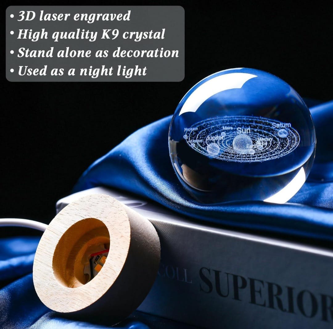 A blue glass crystal ball with a 3D laser-engraved solar system design, displayed on a dark book with a wooden socket and draped blue fabric. A text overlay on the top left lists features: '3D laser engraved', 'High quality K9 crystal', 'Stand alone as decoration', 'Used as a night light'.