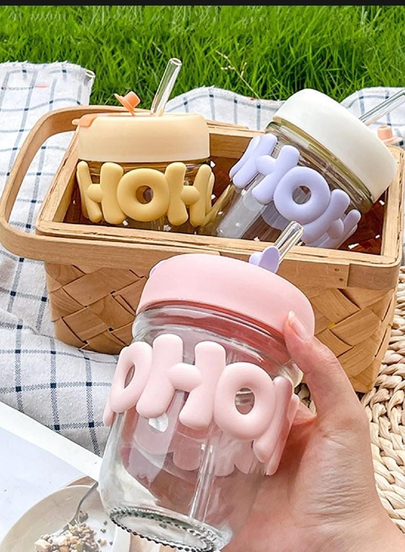 A hand holds a clear glass tumbler with a pink lid and a pink 3D bubble design spelling 'oh my' on the side. In the background, a woven basket holds two more matching tumblers in different pastel colors, set on a checkered blanket with green grass visible in the background.