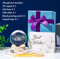 Gift set containing a 3D crystal ball with an LED base, a blue patterned gift box with a purple ribbon, a white greeting card that says 'Best Wishes', a USB cable, and a crystal wipe, displayed on a white surface against a dark blue background with product details listed in the top left.